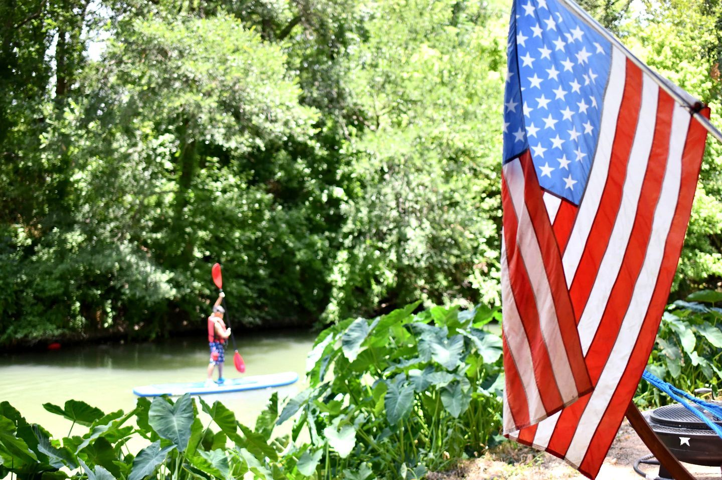 A river with a paddleboarder, surrounded by lush greenery and an American flag.