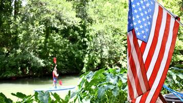 A river with a paddleboarder, surrounded by lush greenery and an American flag.