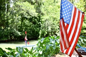 A river with a paddleboarder, surrounded by lush greenery and an American flag. - Amazing Safari Cabin along the Cibolo Creek, Marion, Texas (Marion)