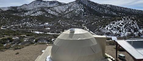 A dome-shaped tent with a solar panel on top, set on a wooden platform with snow patches, surrounded by a mountainous landscape.