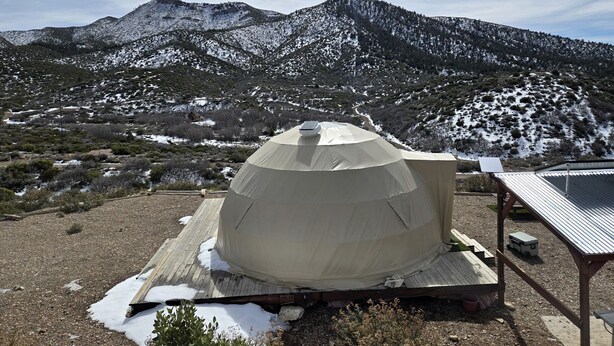 A dome-shaped tent with a solar panel on top, set on a wooden platform with snow patches, surrounded by a mountainous landscape. - Tranquil Desert Retreat in a Geodesic Dome with Scenic Views in Las Vegas, Nevada (Las Vegas)