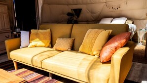 Ein gelbes Sofa mit vielen Kissen, ein StreifenTeppich und im Hintergrund eine zeltartige Konstruktion.