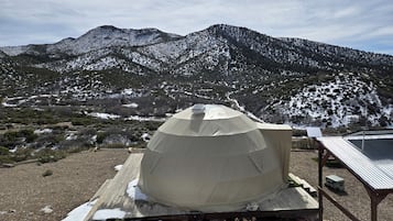 Una tienda de campaña abovedada con un panel solar en la parte superior, ubicada sobre una plataforma de madera con parches de nieve, rodeada por un paisaje montañoso.