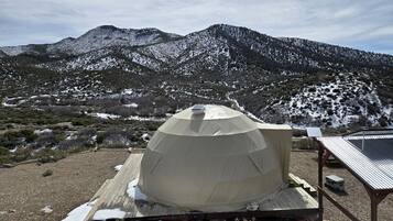 A dome-shaped tent with a solar panel on top, set on a wooden platform with snow patches, surrounded by a mountainous landscape.