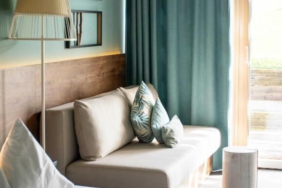 A hotel room with a bed, a sofa, a lamp, a window with curtains, and wall art.