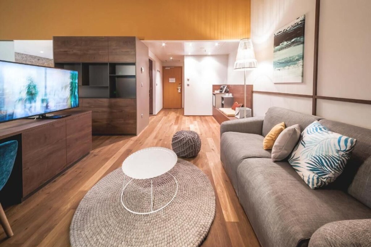 A modern living room with a flat-screen TV, a grey sofa with patterned cushions, a round coffee table, and a wooden floor.