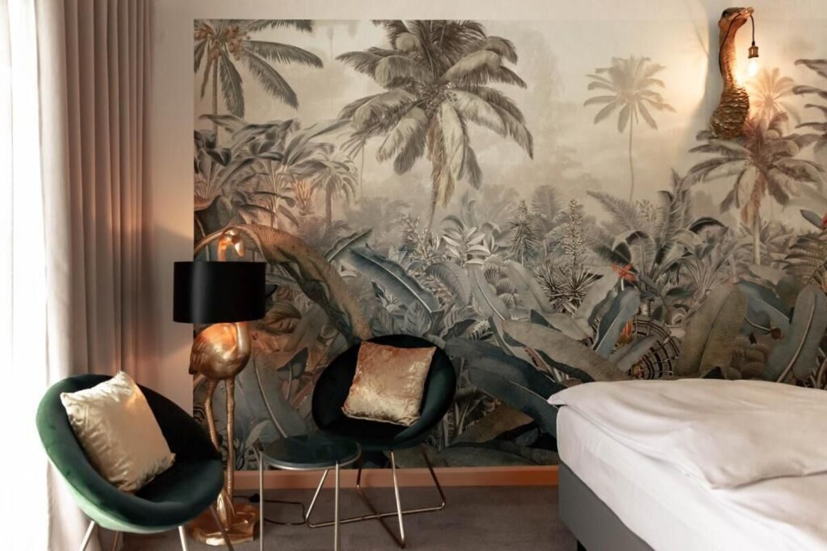 A room with a bed, two chairs, a floor lamp, and a wall mural of tropical foliage.
