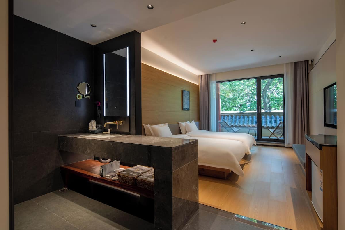 Une chambre d’hôtel moderne dotée d’un grand lit, d’un canapé-lit séparé, d’une coiffeuse avec miroir, d’une petite table et d’un balcon offrant une vue sur les arbres.