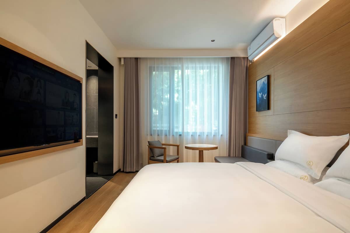 Une chambre d’hôtel avec un grand lit, une petite table ronde, une chaise, un téléviseur fixé au mur et une fenêtre avec des rideaux légers.