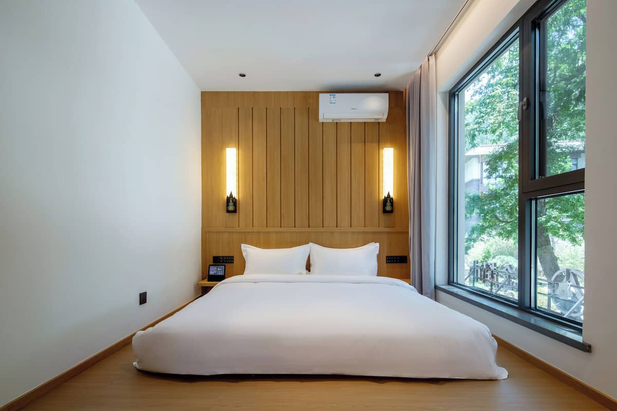 Une chambre d’hôtel moderne dotée d’un grand lit, de murs recouverts de panneaux en bois, de deux lampes murales, d’un climatiseur et d’une grande fenêtre donnant sur un paysage verdoyant.
