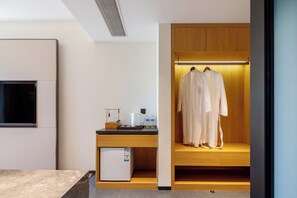 A modern hotel room with a wooden desk, a small refrigerator, a TV, and a walk-in closet with hanging robes. - A Thought Laoshan Cultural Experience (Qingdao)
