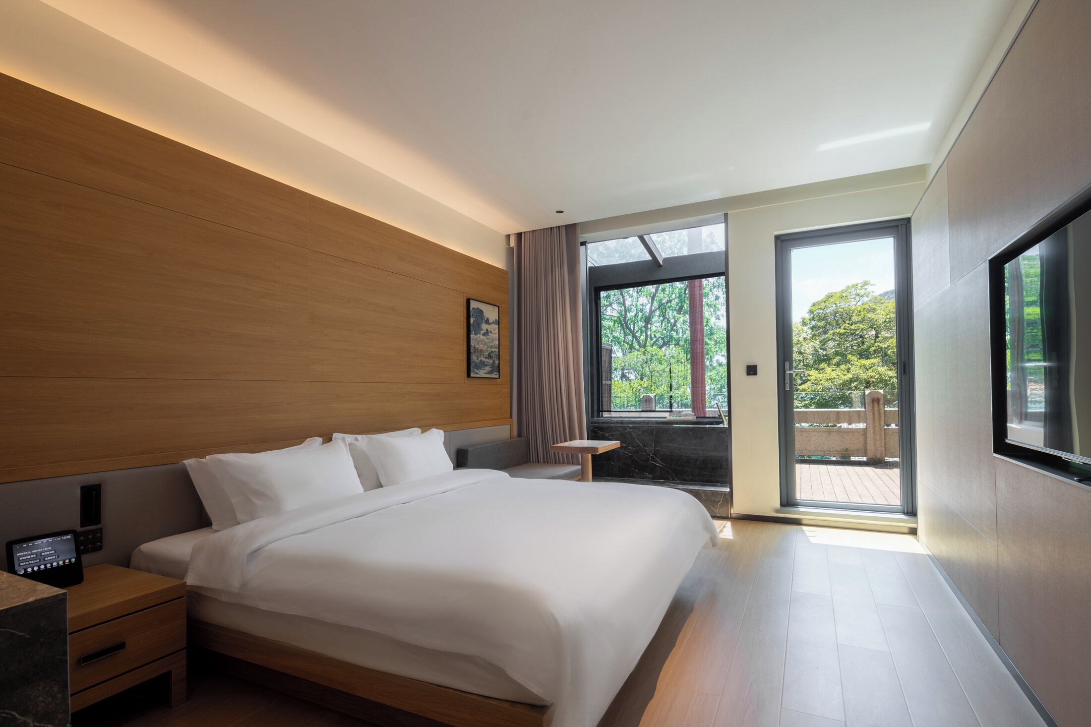 Habitación de hotel moderna con una cama grande, mesita de noche y vistas al verdor por la ventana.