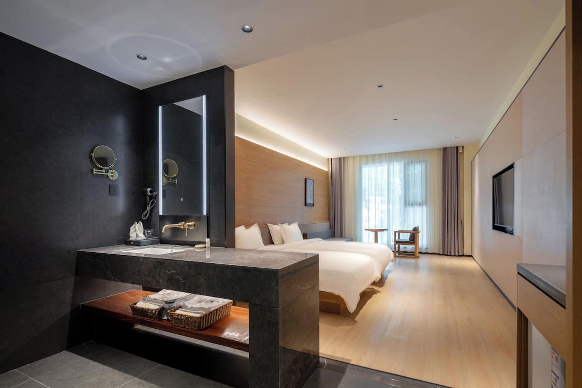 Une chambre d’hôtel moderne dotée d’un meuble-lavabo en marbre, d’un grand lit, d’une table de chevet en bois, d’une télévision et d’une grande fenêtre avec des rideaux.