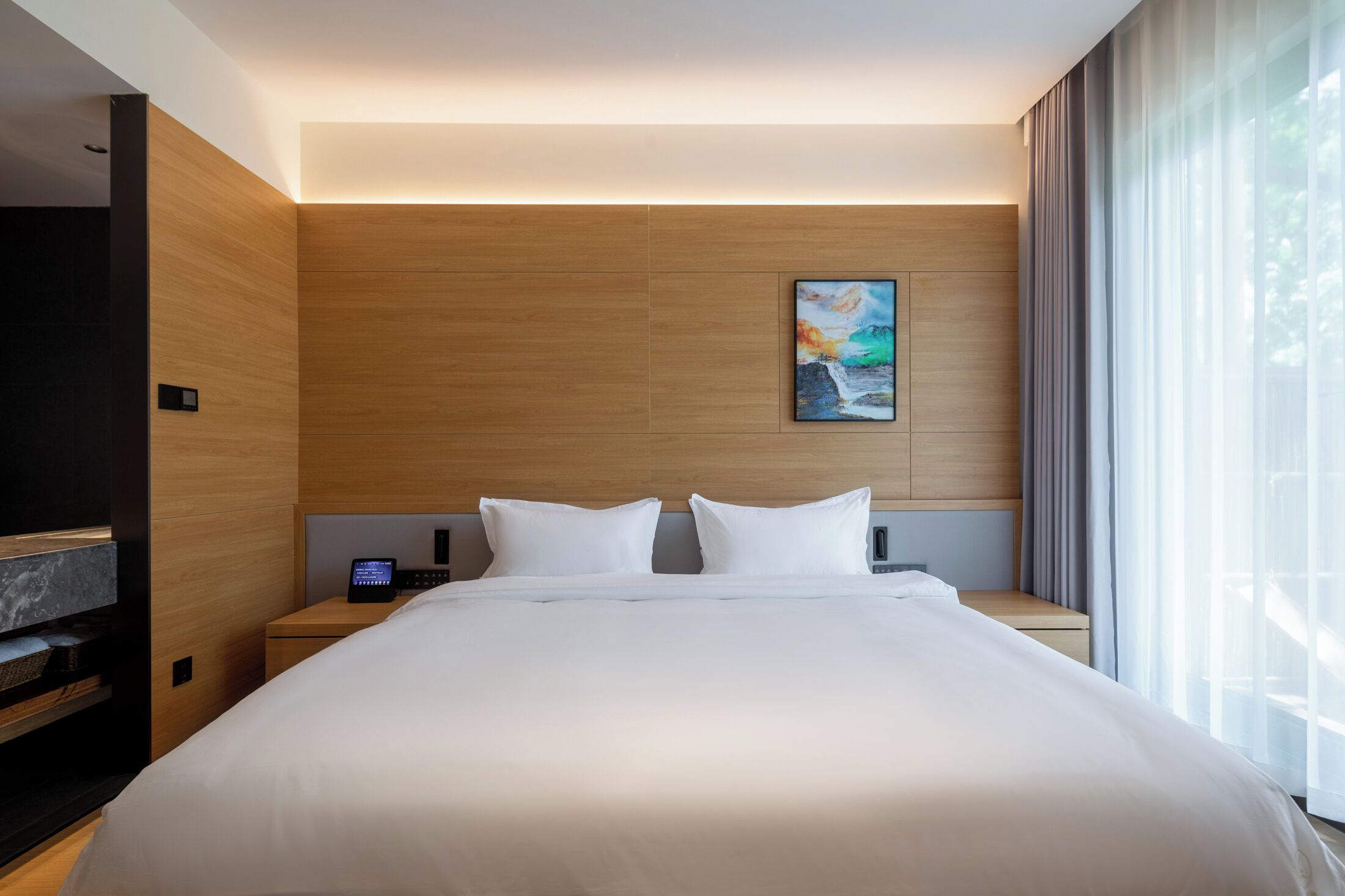 A modern hotel room with a large bed, wooden panel walls, a painting, a bedside table with a phone, and a TV.