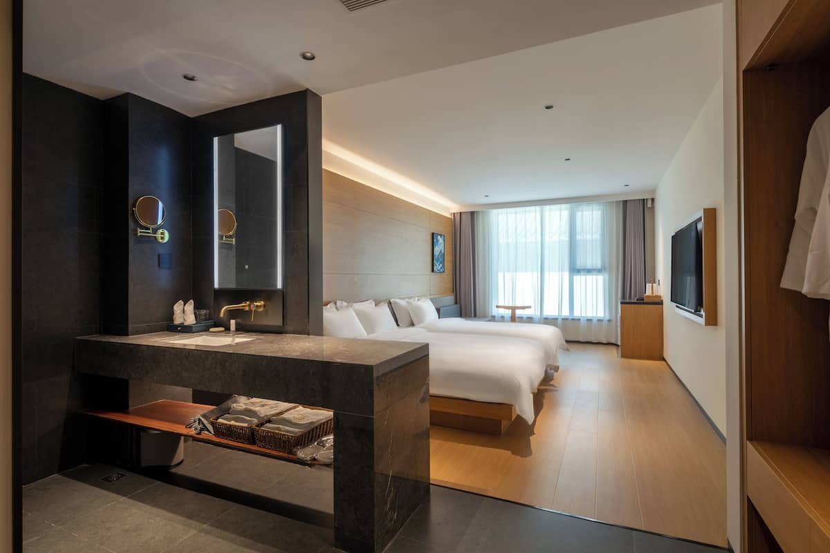 Une chambre d’hôtel moderne dotée d’un grand miroir, d’une coiffeuse en marbre, d’un lit, d’une télévision et d’une fenêtre avec des rideaux.