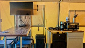 A compact kitchenette with a microwave, coffee maker, and a small refrigerator. There is a table with chairs and a TV mounted on the wall. - Texas Safari Tent Adventures Await in this Lovely San Marcos River Rental (Kingsbury)