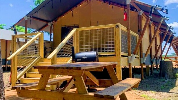 A wooden structure with a picnic table and grill, a fenced area, and a building with a number '49' visible. - Texas Safari Tent Adventures Await in this Lovely San Marcos River Rental (Kingsbury)