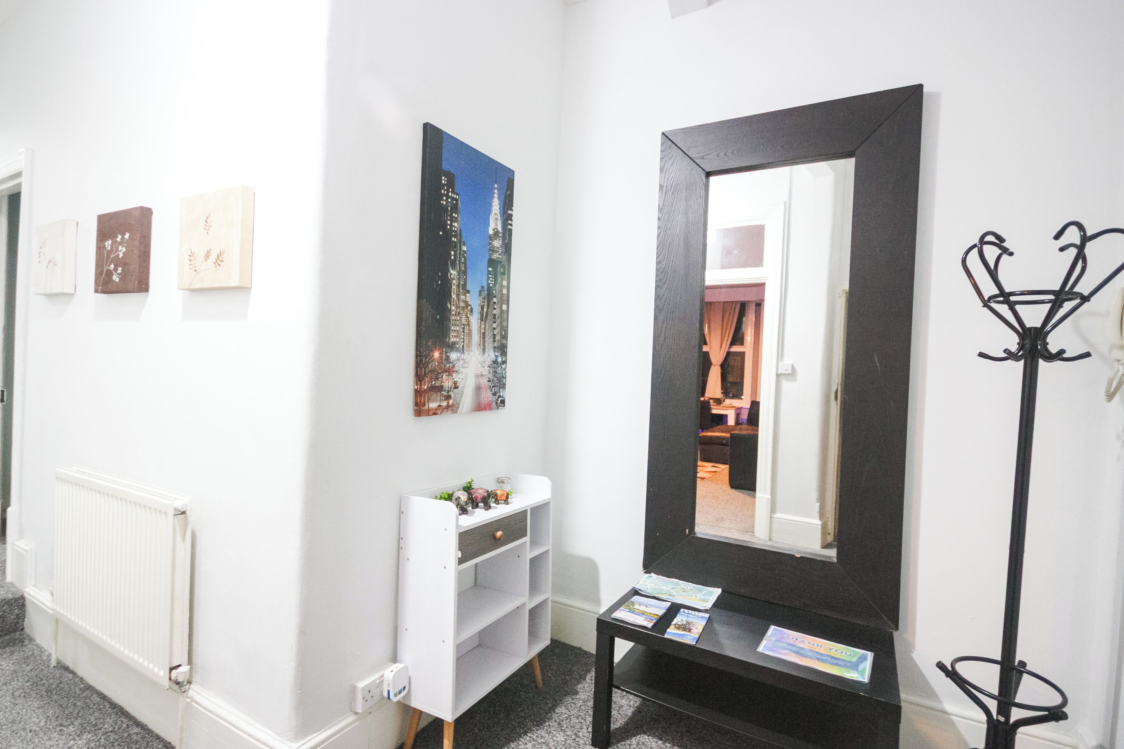 A hotel lobby with a white wall, a black-framed mirror, a coat rack, and a small table with a magazine.