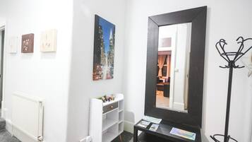 A hotel lobby with a white wall, a black-framed mirror, a coat rack, and a small table with a magazine.
