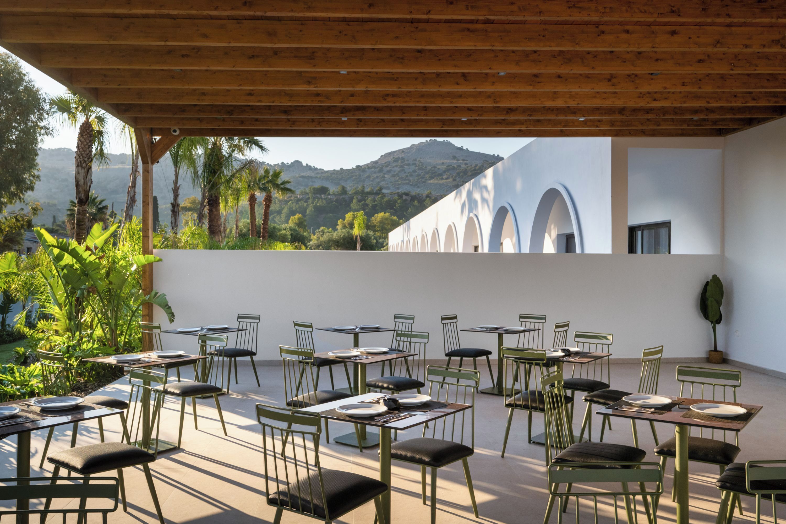 An outdoor dining area with tables and chairs, surrounded by greenery and a view of a mountain.