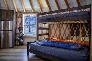 A cozy cabin interior with a bunk bed, a refrigerator, a bookshelf, and a window with a snowy view.