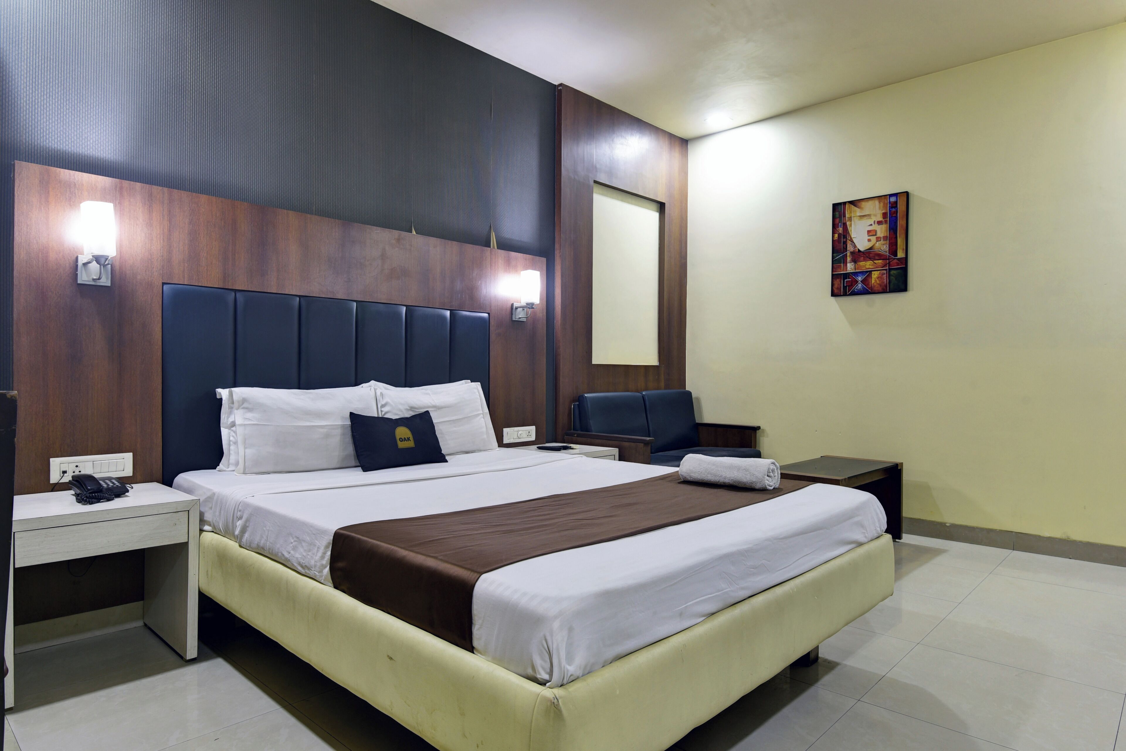 Ein Hotelzimmer mit einem großen Bett, Nachttischen, einem Schreibtisch, einem Sessel und einem an der Wand hängenden Kunstwerk.