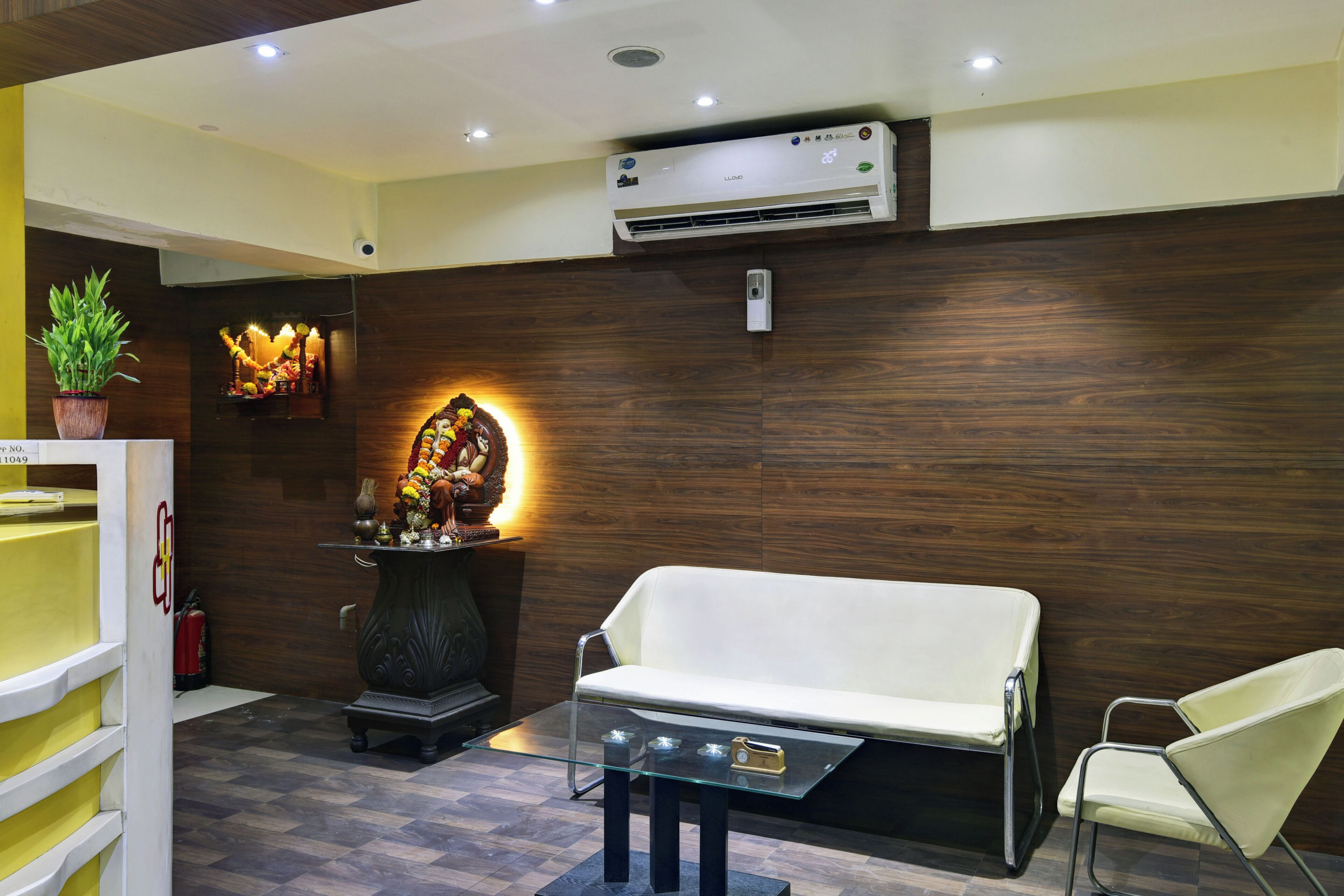 A reception area with a sofa, a glass table, a potted plant, a wall-mounted air conditioner, and a decorative wall piece.