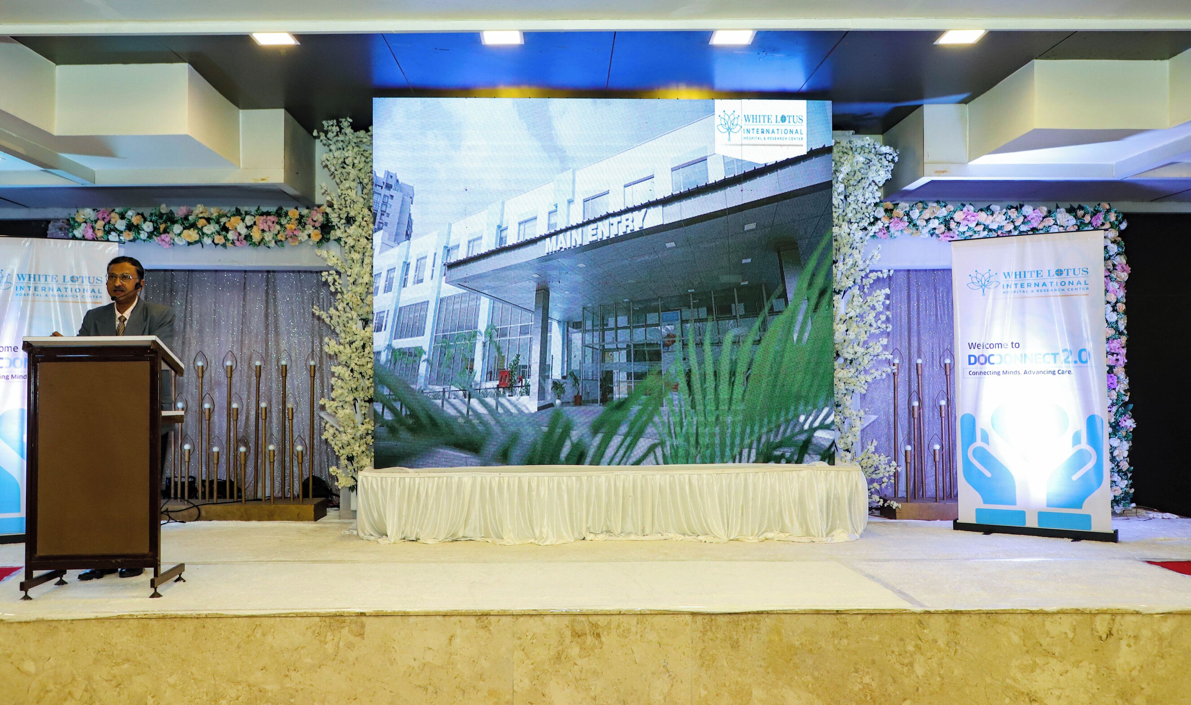 Eine Bühne mit Rednerpult, einer großen Leinwand, die ein Gebäude zeigt, und Blumendekorationen.