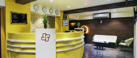 A hotel reception area with a yellow counter, a reception desk, a clock, and a wall-mounted air conditioning unit.