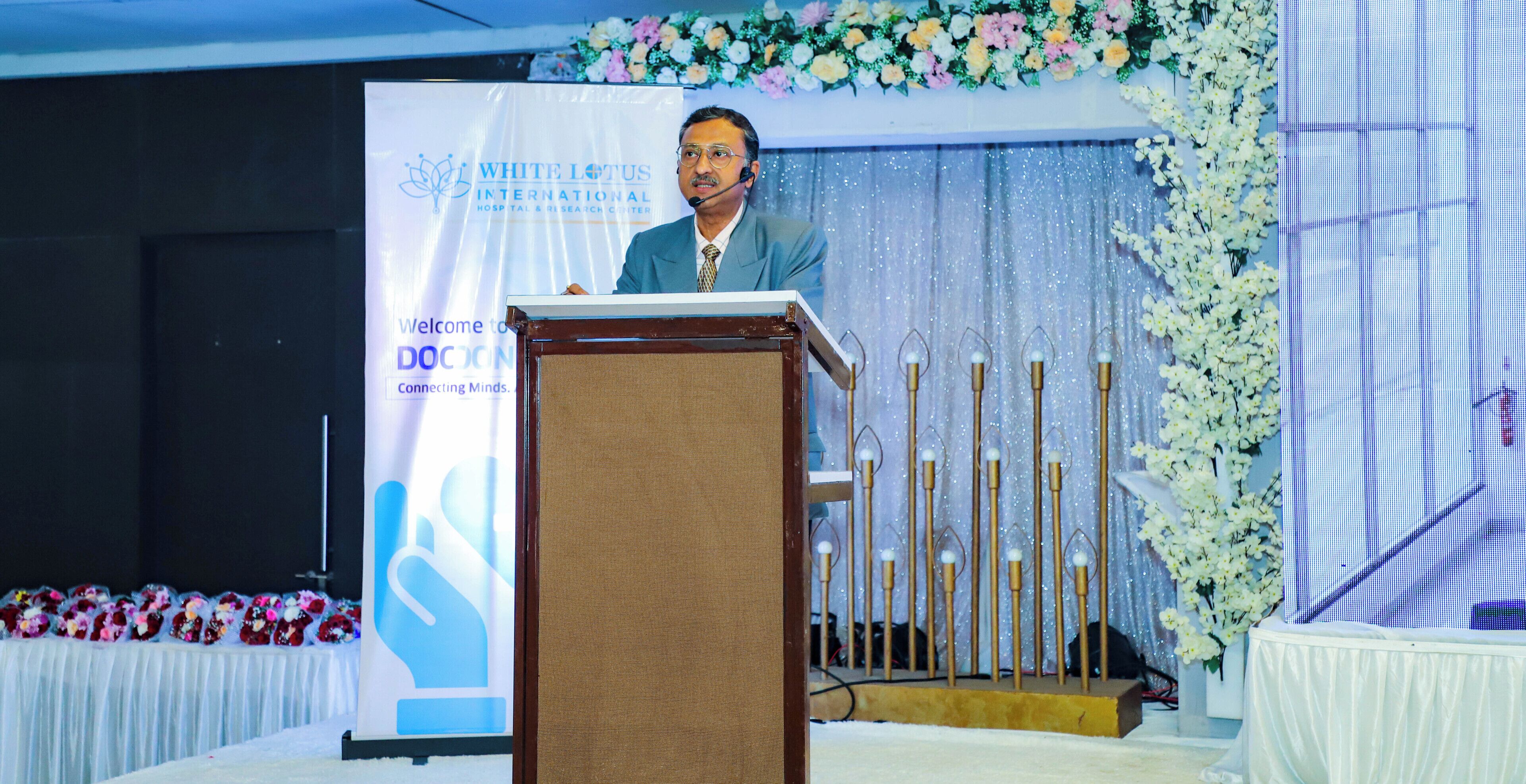 A speaker at a podium with a backdrop displaying 'WHITE LOTUS INTERNATIONAL HOSPITAL & RESEARCH CENTER' and 'DOCON Connecting Minds.'