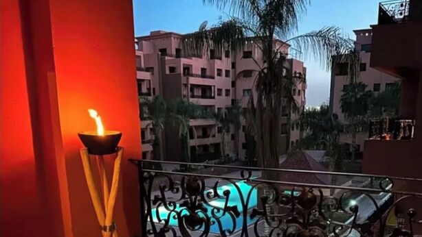 Ein Balkon mit zwei schwarzen Stühlen, einem kleinen runden Tisch, einer dekorativen Lampe und Blick auf Palmen und Gebäude.