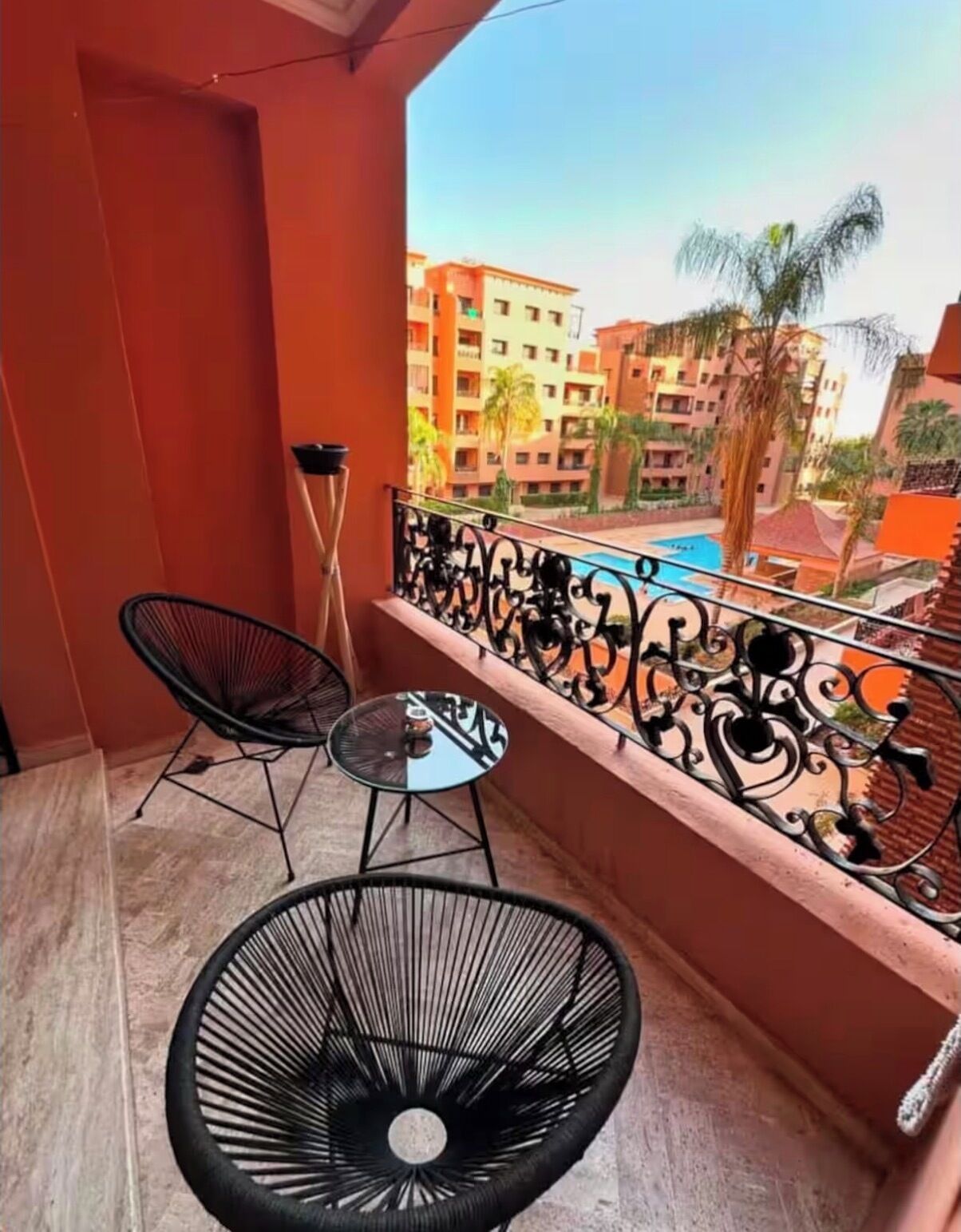 A balcony with a table and two chairs, a view of a swimming pool, and surrounding buildings.