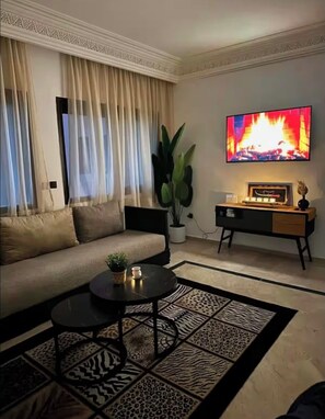 Ein Wohnzimmer mit einer Couch, einem Couchtisch, einem Fernseher der ein Feuer zeigt, einer Pflanze und einem Beistelltisch.