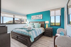 Ein Schlafzimmer mit einem Bett, Nachttischen, einem Sessel, einer Kommode und einer Wand mit Blick auf Gebäude.