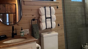 A bathroom with wooden walls, a window, a mirror, a sink, a toilet, and a shower area.