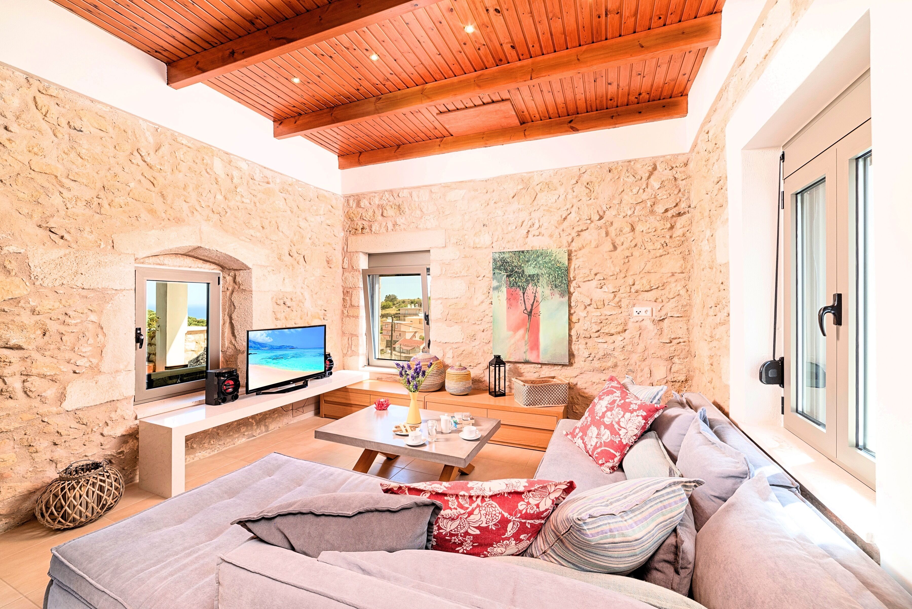 A cozy living room with stone walls, a wooden ceiling, a large sofa, a TV, and a window with a view.