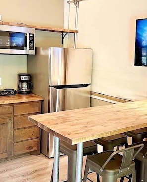 Eine kleine Küche mit Mikrowelle, Kühlschrank, Kaffeemaschine und einem Holztisch.