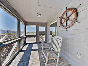 Eine überdachte Terrasse mit Blick auf den Strand, ein Schiffsrad und eine Reihe weißer Stühle.
