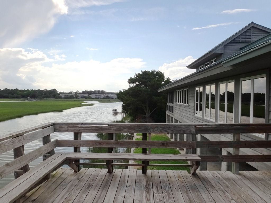 A wooden deck with a bench overlooking a river, a building with large windows, and a grassy area with trees.