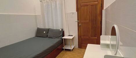 A small, single-bed room with a wooden door, a small bedside table, a mirror, and a white dresser.