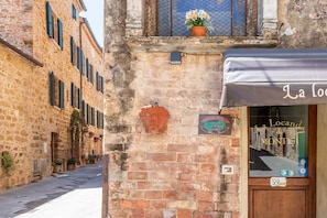 Una calle estrecha con edificios de piedra, una tienda con el rótulo 'La Locand di Montisi' y una planta en el alféizar.