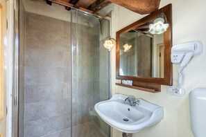 Ein Badezimmer mit Duschkabine aus Glas, einem weißen Waschbecken, einem Spiegel mit Holzrahmen und einem an der Wand befestigten Föhn.