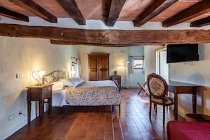 A bedroom with a wooden ceiling, a bed with a floral bedspread, a nightstand, a chair, and a television mounted on the wall. - Locanda di Montisi Torre Room among the Montalcino Hills (Montisi)