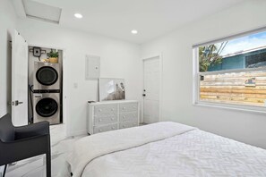 A bedroom with a bed, a dresser, a washing machine, and a window with a view of a wooden fence. - Marina Row 1 - Near Atlantic Ave (Delray Beach)