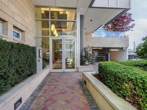 Ein Gebäudeeingang mit Glastür, einem gepflasterten Weg und einer Hecke auf der rechten Seite.