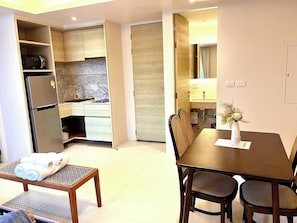 A modern kitchen with a built-in microwave, a refrigerator, and a sink. There is a dining area with a table and chairs, and a small bench with towels. - Adventure Condo Aonang (Krabi)