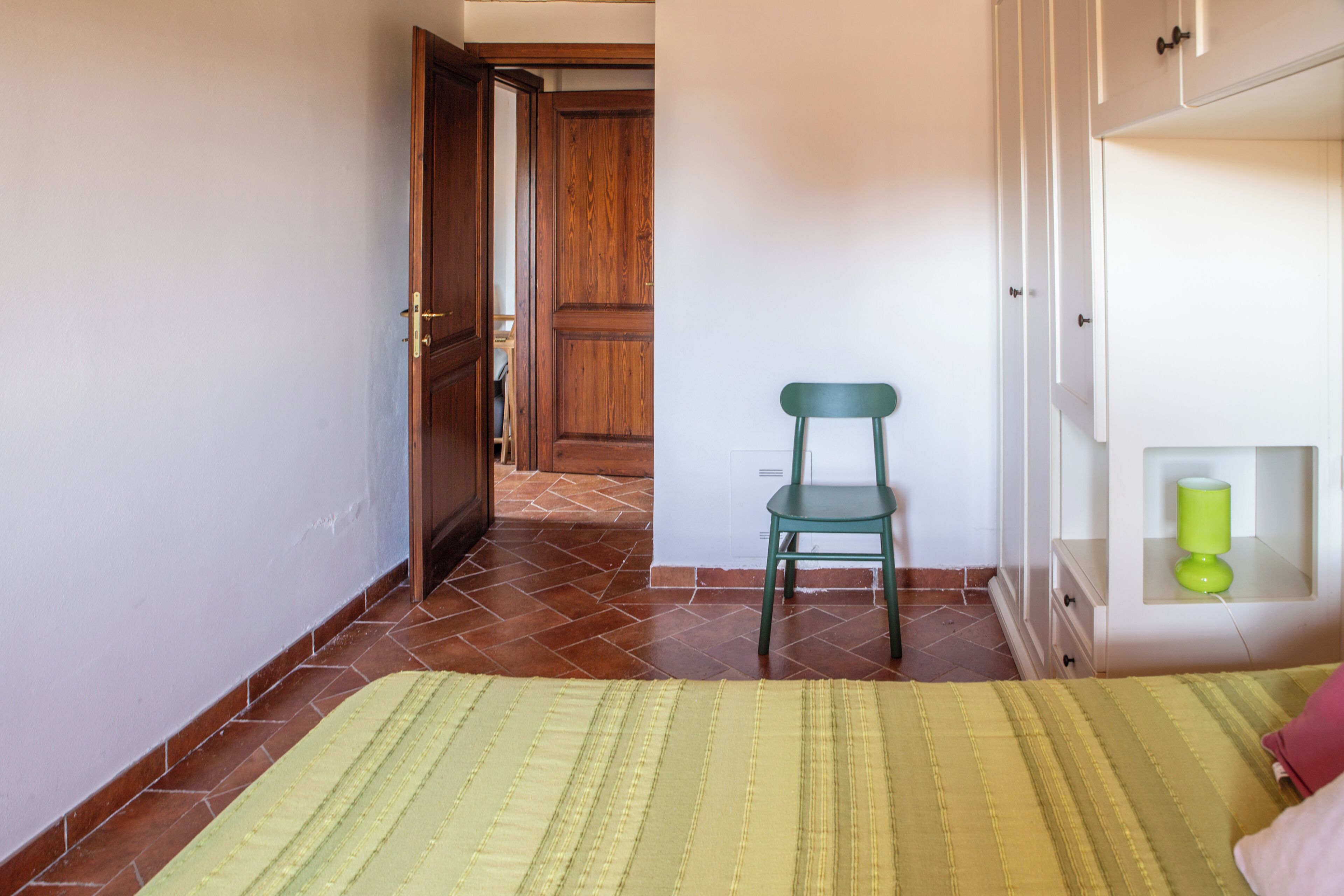 Una habitación con una cama, una silla, una puerta de madera y un armario blanco con una lámpara verde.