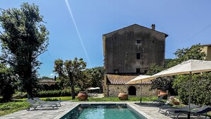 A rectangular swimming pool with clear water, surrounded by a tiled deck. There are lounge chairs and umbrellas, and a large building with a tiled roof in the background. - Borgo San Jacopo - Il Cipressino (Vicopisano)