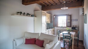 A living room with a white sofa, a red pillow, a wooden ceiling, a kitchen area with a white refrigerator, a dining table with chairs, and a window with a view. - Borgo San Jacopo - Il Cipressino (Vicopisano)