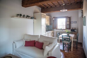 A living room with a white sofa, a red pillow, a wooden ceiling, a kitchen area with a white refrigerator, a dining table with chairs, and a window with a view. - Borgo San Jacopo - Il Cipressino (Vicopisano)